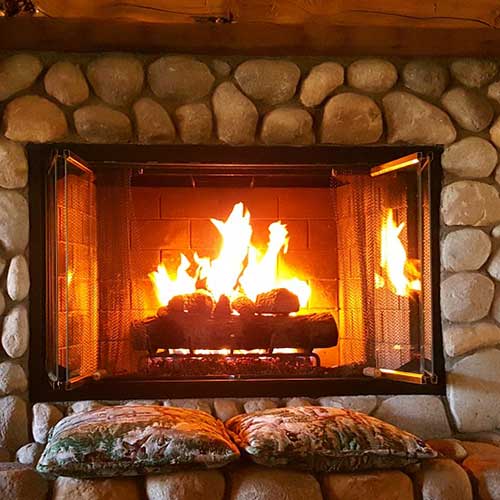 Wood fireplace with burning logs with doors and large round river rock surround - pillows to sit on