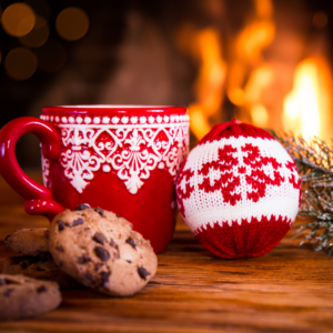red festive mug next to a red and white knitted ornament and some cookies with a fire burning in the background